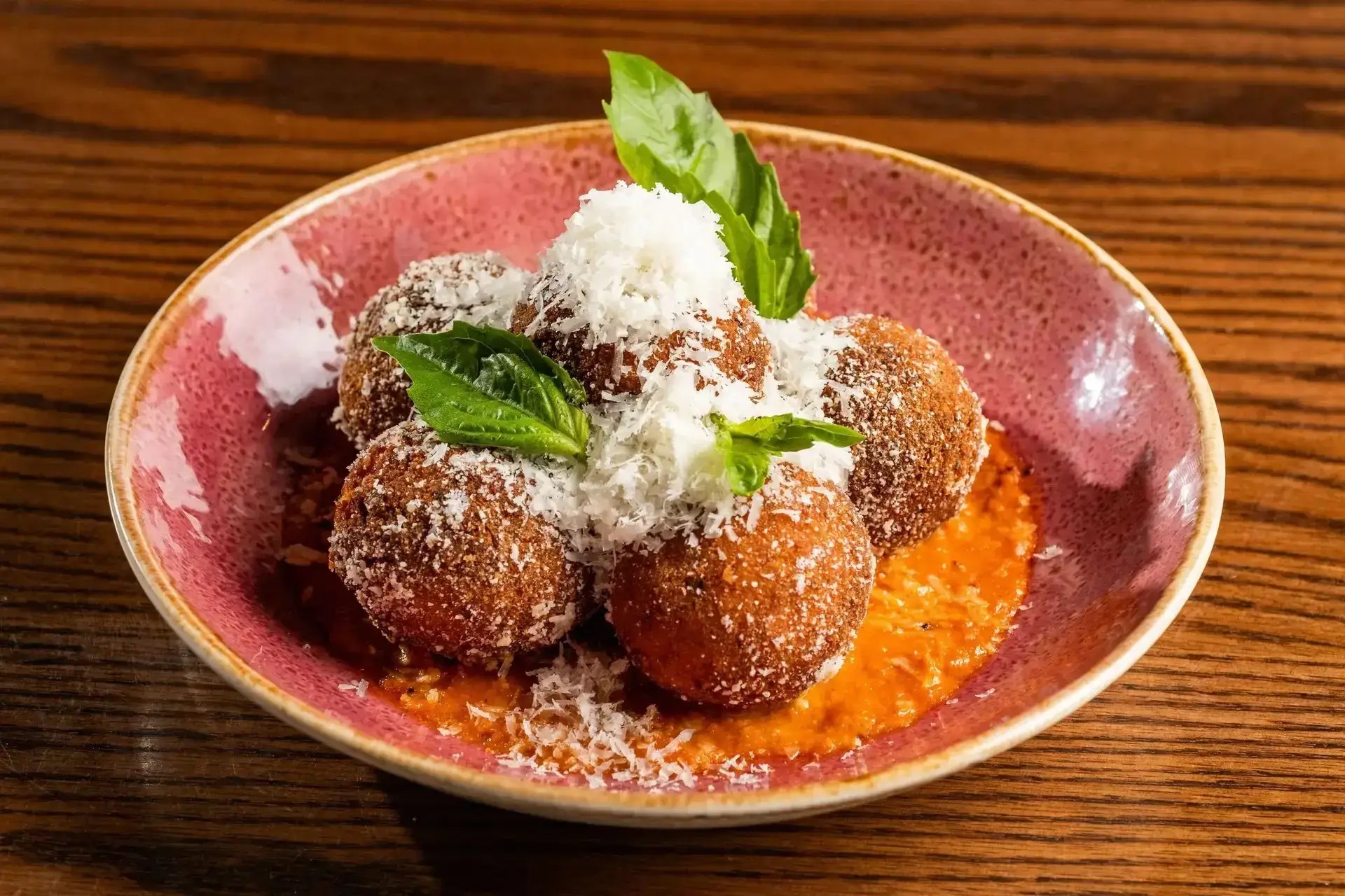 Golden-brown crispy Italian arancini rice balls served over marinara sauce, topped with grated parmesan cheese and fresh basil leaves in a pink bowl.