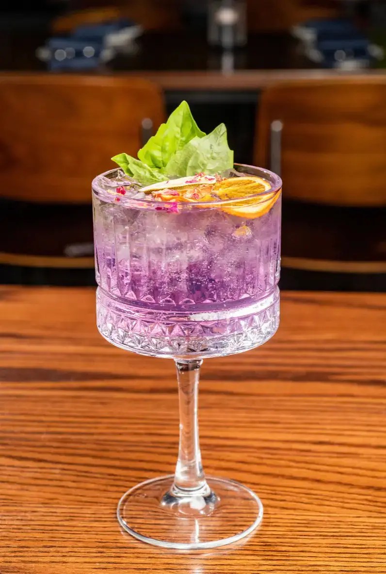 An elegant purple craft cocktail served in a textured crystal goblet glass, garnished with a dried orange slice, fresh basil leaves, and flower petals on a wooden table.