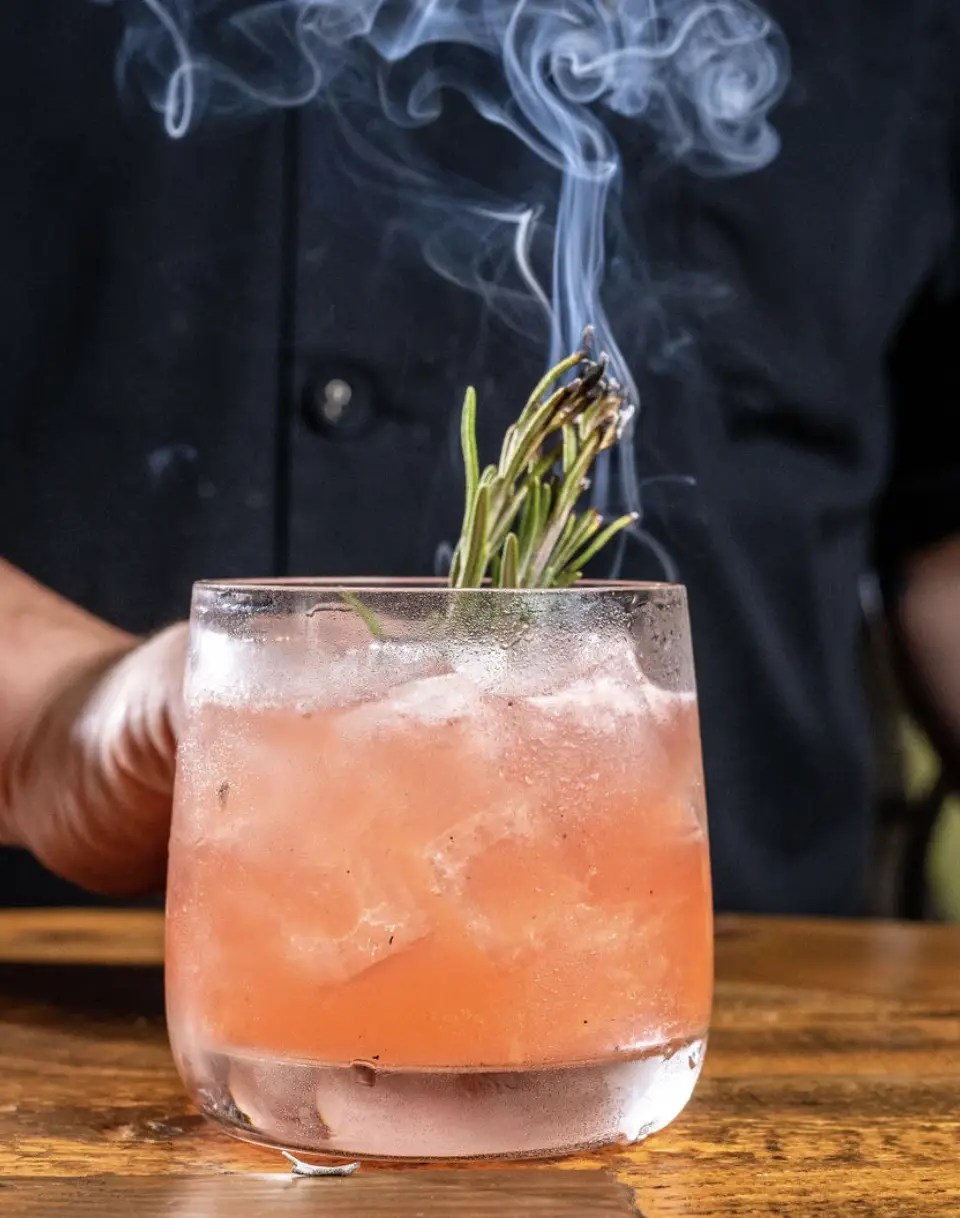 A dramatic close-up of a pink craft cocktail served with crushed ice and a burning sprig of smoked rosemary releasing swirls of smoke, held by a bartender.